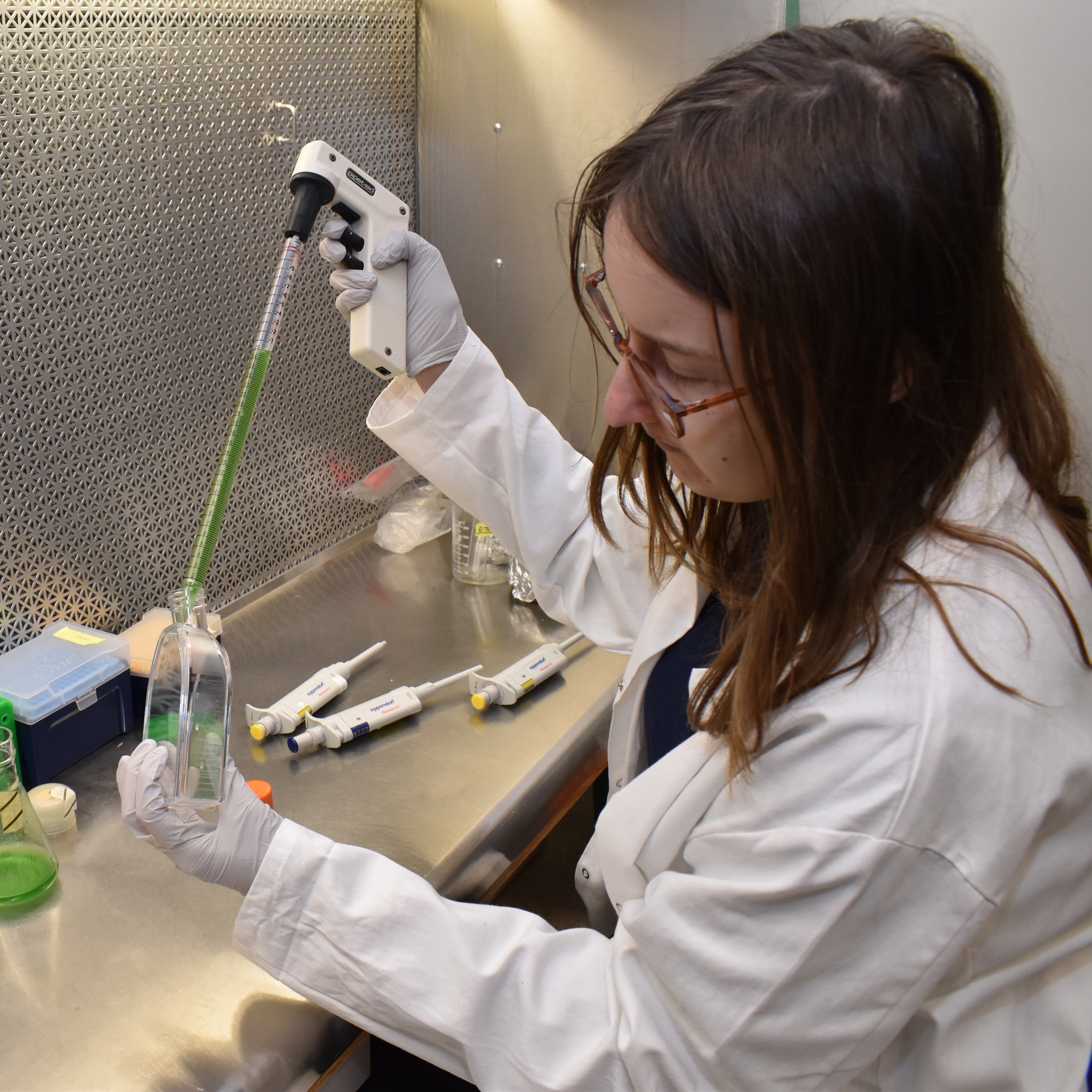 A person in a lab coat uses a pipette to move green liquid to a scientific flask.